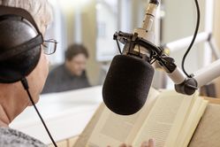 Foto einer Szene aus dem Hörbuchstudio: Die Sprecherin sitzt vor einem Mikrofon an einem Tisch und liest aus einem Buch vor. Im Hintergrund hinter einer Glasscheibe sitzt der Aufnahmeleiter.