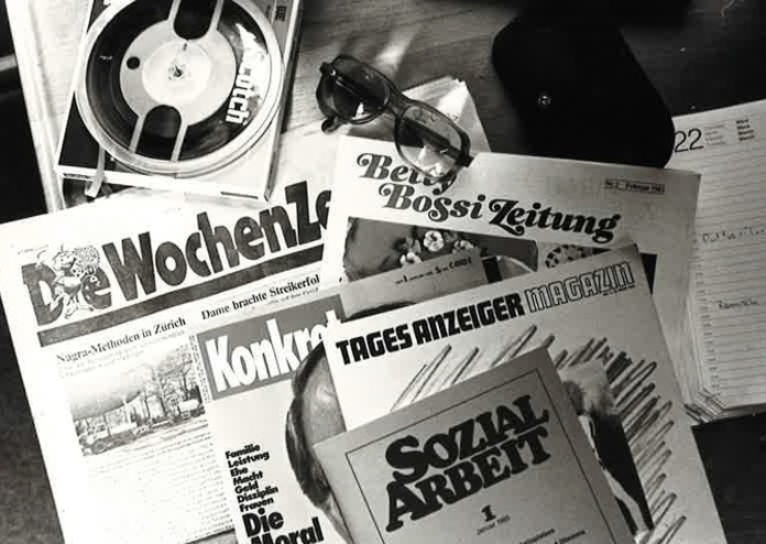 Abbildung eines Tonband-Abspielgeräts sowie einigen Zeitschriften wie zum Beispiel «Die Wochenzeitung»,  «Betti Bossi Zeitung» und «Sozialarbeit». 